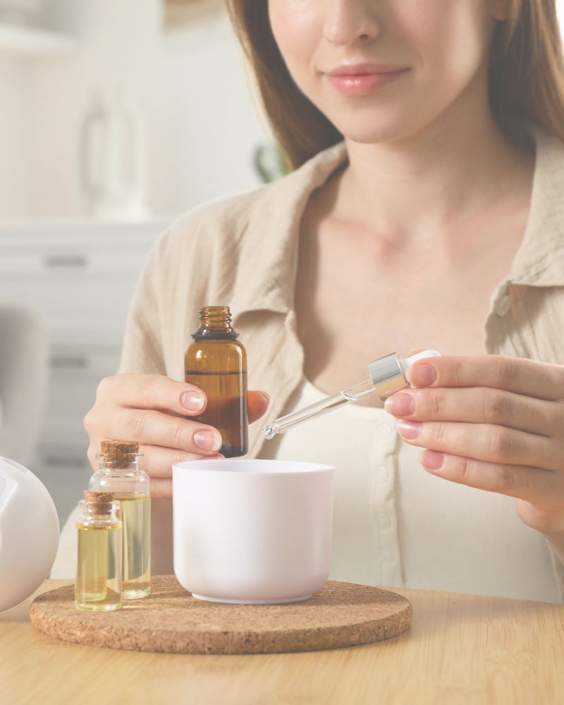 Mulher em momento de autocuidado utilizando óleo essencial Swaminah em difusor de ambiente, foco nas mãos e no frasco, estética clean e sofisticada.