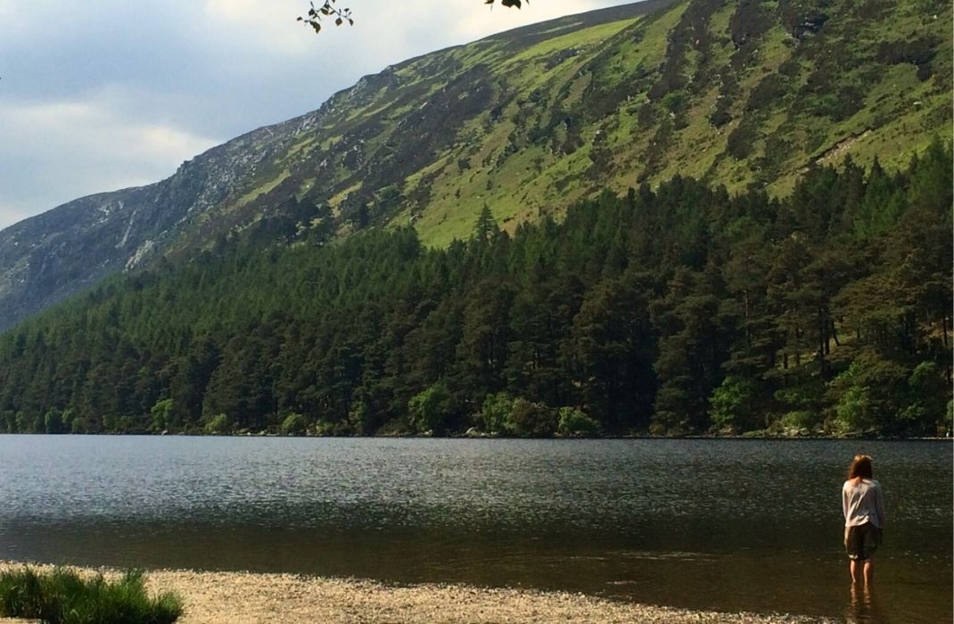 Fundadora da Swaminah de costas diante de um lago e montanhas na Irlanda, em um momento de profunda conexão espiritual e descoberta.